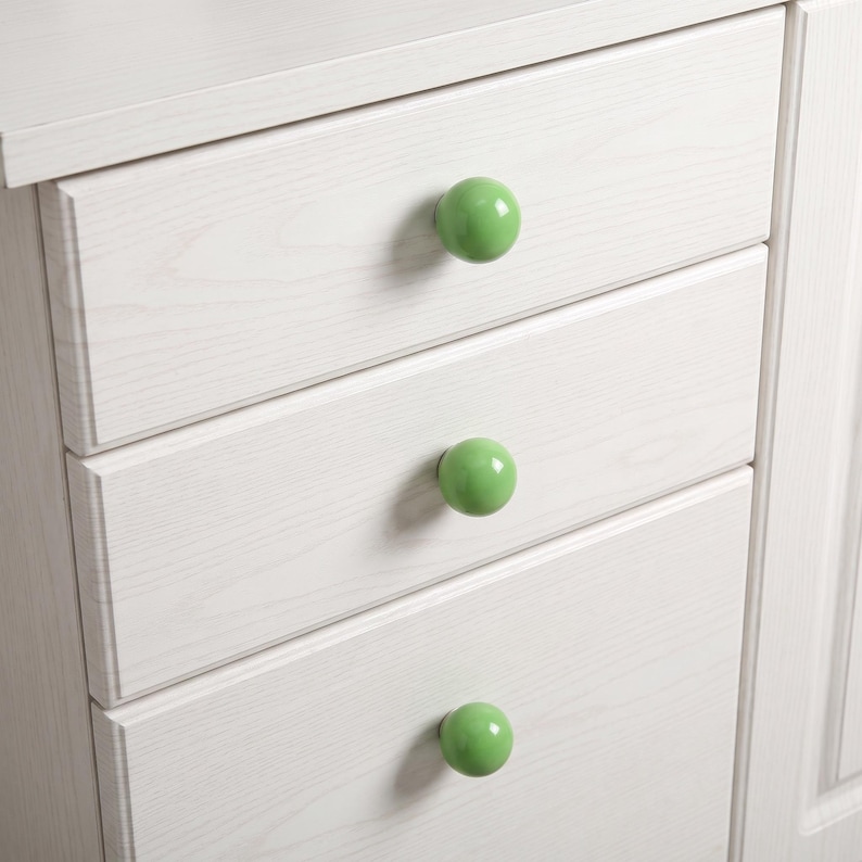 Puede incluir: Una c&oacute;moda de madera blanca con tres cajones apilados, cada uno equipado con un pomo redondo, liso y de color verde claro. La veta de la madera es visible en los frentes de los cajones, aportando una apariencia texturizada al mueble.