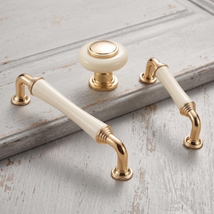 May include: A set of cabinet hardware including one round knob and two arched drawer pulls. Each piece features a smooth, cream-colored cylindrical or circular center section accented by polished gold-tone metal ends and mounting bases.