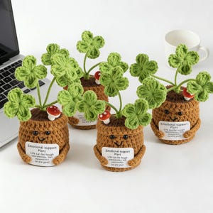 Puede incluir: Plantas de apoyo emocional tejidas a ganchillo con hojas de trébol verdes, pequeños acentos de setas rojas y blancas y caras sonrientes. Cada planta está en una maceta marrón con una etiqueta que dice "Emotional support Plant".