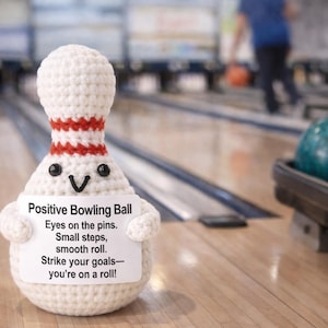 May include: A crocheted bowling pin with a smiling face and arms, wearing a red and white striped collar. The pin has a sign that reads "Positive Bowling Ball" with motivational text. Bowling balls are in the background.
