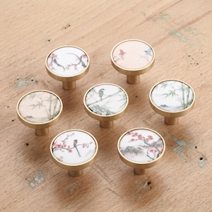 May include: Seven round cabinet knobs with a brushed gold metal base and a white ceramic top. Each knob features a unique hand-painted design including birds, bamboo stalks, and flowering branches in shades of red, green, and black ink.