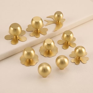 Può includere: Una collezione di nove pomelli per armadietti in metallo color oro, ciascuno dotato di una maniglia sferica. I pomelli sono montati su varie piastre decorative, incluse forme che ricordano fiori, cerchi smerlati e una sagoma di uccello.
