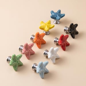Puede incluir: Una colección de nueve pomos de cajón de cerámica con forma de estrella de mar, cada uno con una superficie texturizada con pequeños puntos en relieve. Los pomos vienen en varios colores: azul, amarillo, naranja, rojo, rosa, blanco, verde, azul claro y negro.
