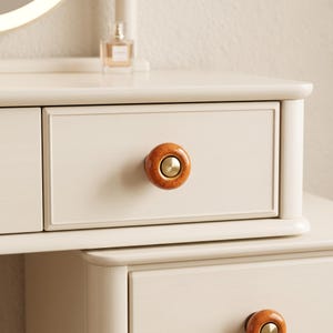 May include: A cream-colored wooden vanity table featuring two drawers with rounded, speckled orange ceramic knobs. Each knob has a smooth, circular brass-toned metal center insert. The vanity surface is smooth and clean with a light finish.