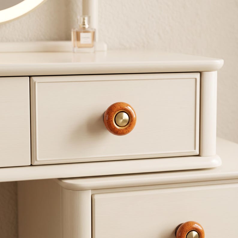 May include: A cream-colored wooden vanity table featuring two drawers with rounded, speckled orange ceramic knobs. Each knob has a smooth, circular brass-toned metal center insert. The vanity surface is smooth and clean with a light finish.