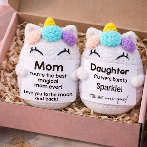 May include: Two white crochet unicorn dolls with closed eyes, a yellow horn, and colourful accents, presented in a gift box. One doll says "Mom" and the other says "Daughter" with sweet messages.