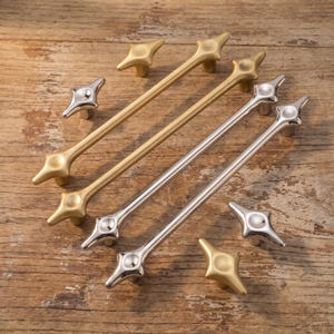 May include: A collection of cabinet hardware including two long drawer pulls and four matching knobs. Two pieces feature a brushed gold finish, while the other four pieces have a polished silver finish. Each item has a four-pointed star shape.