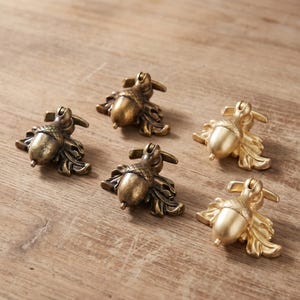 May include: Five metal drawer pulls shaped like acorns with attached leaves. Three pulls feature a dark, antiqued bronze finish with visible texture, while two pulls have a smooth, matte gold finish. Each pull includes a hinged top mounting bracket.