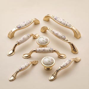 May include: A collection of gold-toned metal cabinet hardware including several arched drawer pulls and two round knobs. Each piece features a central ceramic inlay decorated with a delicate floral pattern in shades of purple, green, and brown.