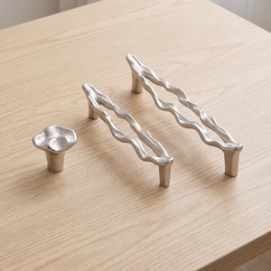 Könnte beinhalten: Ein Set aus silberfarbenen Möbelbeschlägen, bestehend aus einem kleinen, runden Knopf mit unregelmäßiger, organischer Form und zwei länglichen Schubladengriffen mit wellenförmigem, offenem Rahmendesign, glatter Oberfläche und zwei Stützen.