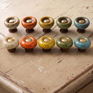 May include: Ten round ceramic cabinet knobs arranged in two rows of five. Each knob features a glossy, speckled glaze in various colors including beige, orange, yellow, green, and blue, with a central circular brass-toned metal inlay.