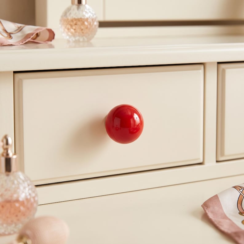 Puede incluir: Un pomo de caj&oacute;n esf&eacute;rico, brillante y de color rojo intenso, centrado en un frente de caj&oacute;n de madera de color crema. El pomo presenta una superficie lisa y reflectante que contrasta con el acabado mate del mueble circundante.