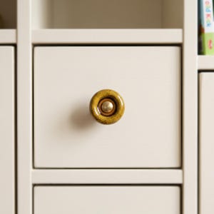 May include: A circular cabinet knob featuring a speckled mustard yellow ceramic finish with a smooth, recessed center. The center of the knob contains a small, polished brass-toned metal disc, providing a contrast to the textured ceramic surface.