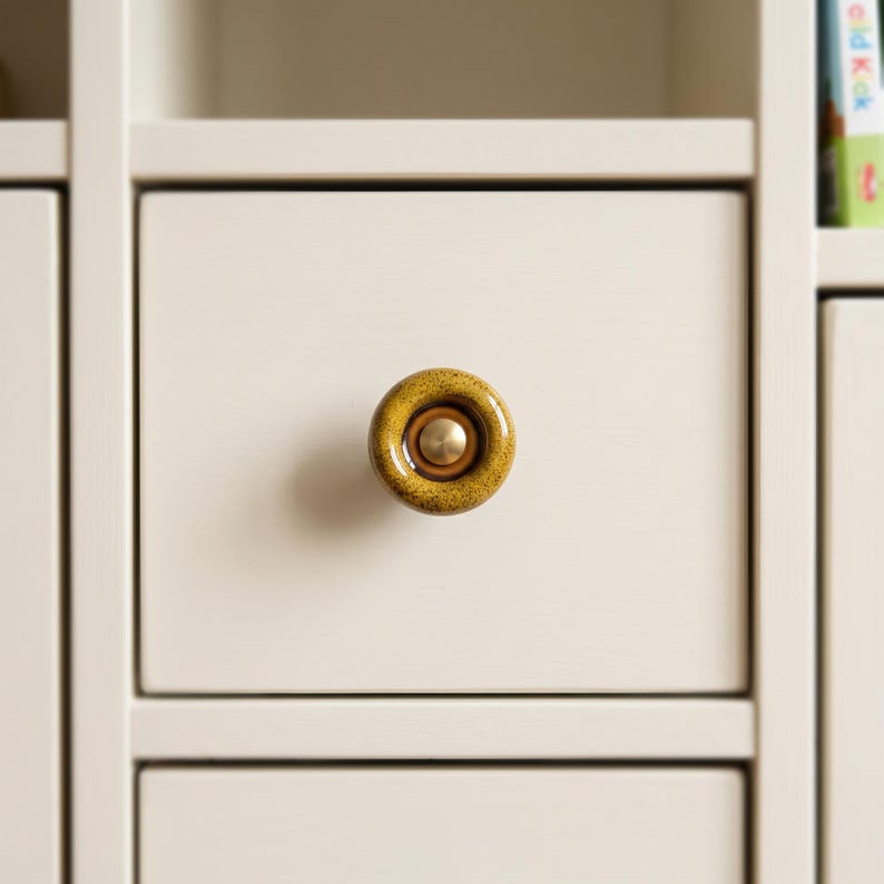 May include: A circular cabinet knob featuring a speckled mustard yellow ceramic finish with a smooth, recessed center. The center of the knob contains a small, polished brass-toned metal disc, providing a contrast to the textured ceramic surface.