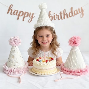 May include: A young girl smiles, wearing a white birthday hat with lace and a pom-pom. A cake with candles sits in front of her, and two other party hats are on either side. A banner that says "happy birthday" hangs in the background.