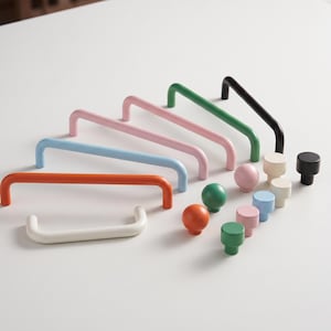 May include: A collection of cabinet hardware including several U-shaped drawer pulls and various round knobs in a spectrum of colours including orange, light blue, pink, green, black, and white, arranged in a group on a plain surface.