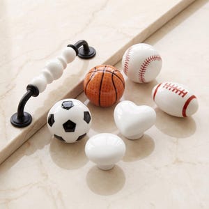 May include: A collection of cabinet hardware including one long white and black handle and six knobs shaped like a football, a basketball, a baseball, an American football, a heart, and a plain white sphere, all featuring smooth ceramic finishes.