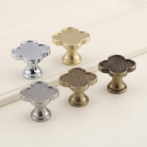 May include: Five clover-shaped cabinet knobs with a grid-textured top surface. The set includes one polished chrome knob, one brushed nickel knob, two antique brass knobs, and one dark bronze knob, each featuring a central pedestal base.