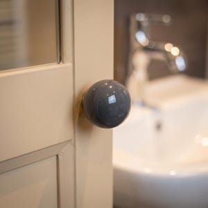 Puede incluir: Un pomo de armario esf&eacute;rico, liso y de color gris oscuro brillante, fijado a un marco de puerta de madera de color crema claro. El pomo presenta una superficie reflectante que capta la luz, situado frente a un fondo borroso de lavabo y grifo.