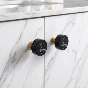 May include: Two circular cabinet knobs featuring a polished black marble face with white veining, mounted on a brushed brass base. These hardware pieces are attached to white marble cabinet doors with grey veining, creating a high-contrast look.