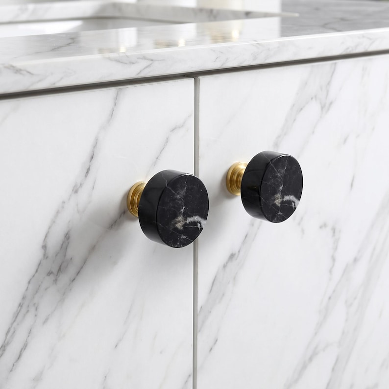 May include: Two circular cabinet knobs featuring a polished black marble face with white veining, mounted on a brushed brass base. These hardware pieces are attached to white marble cabinet doors with grey veining, creating a high-contrast look.
