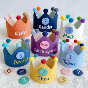 May include: A collection of colorful felt crowns, each with a unique name and the number 3. The crowns are in shades of orange, blue, white, purple, yellow, and light blue, adorned with pom-poms. Names include Atlas, Xander, Malik, Amelia, Theo, Rosie, and Tobias.