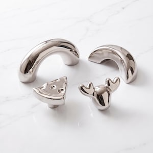 May include: A set of four silver-toned metal cabinet knobs and pulls. The collection includes two smooth, arched handles of varying sizes, one knob shaped like a slice of pizza, and one knob shaped like the head of a moose with prominent antlers.