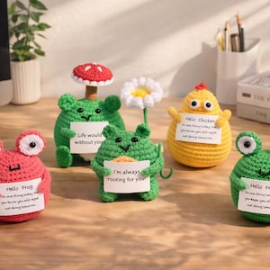 Pode incluir: Uma coleção de figuras de crochê feitas à mão, incluindo sapos, um pintinho e um cogumelo. Cada figura segura uma pequena placa com uma mensagem. As figuras são de várias cores, incluindo verde, vermelho e amarelo.