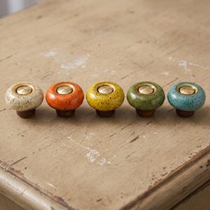 May include: Five round cabinet knobs arranged in a row, each featuring a glossy, speckled ceramic top in cream, orange, yellow, green, and blue. Each knob has a central circular brass-toned metal inlay and a dark brown wooden base attachment.