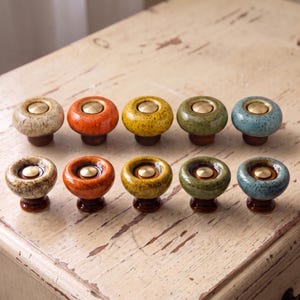 May include: A collection of ten round ceramic cabinet knobs arranged in two rows. Each knob features a glossy, speckled glaze in various colors including beige, orange, yellow, green, and blue, with a central circular brass-toned metal inlay.