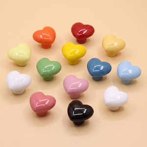 May include: A collection of thirteen heart-shaped cabinet knobs in a variety of glossy colors including red, orange, yellow, green, blue, pink, white, and black. Each knob features a smooth, rounded surface and a central mounting point underneath.