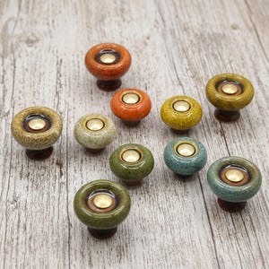 May include: A collection of nine round ceramic cabinet knobs in various colors including orange, yellow, green, and blue. Each knob features a smooth, glossy finish with a speckled texture and a central circular brass-toned metal inlay.