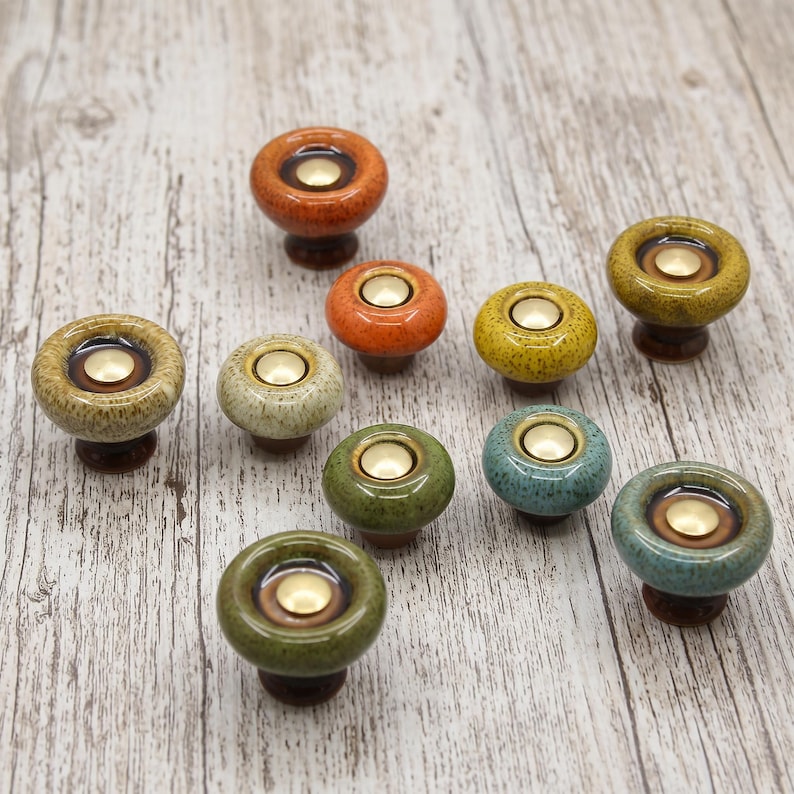May include: A collection of nine round ceramic cabinet knobs in various colors including orange, yellow, green, and blue. Each knob features a smooth, glossy finish with a speckled texture and a central circular brass-toned metal inlay.
