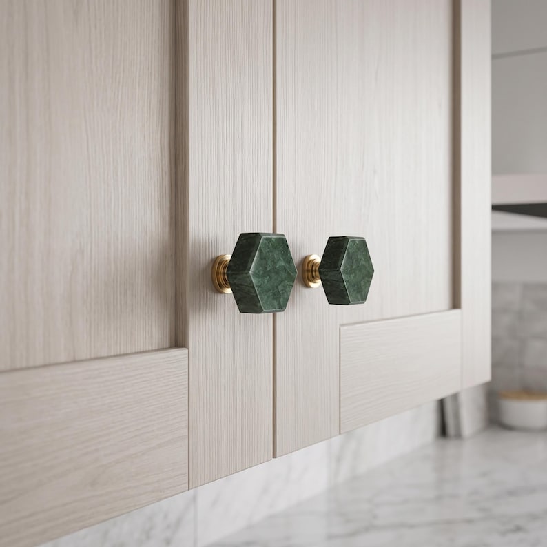 May include: Two hexagonal cabinet knobs made of dark green marble with natural veining, each mounted on a circular brass base. These knobs are attached to light-coloured, wood-grain cabinet doors with a vertical panel design and clean lines.