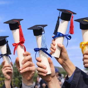 May include: Hands holding graduation diplomas topped with black mortarboards and colorful tassels. Each diploma is tied with a ribbon in red, blue, and yellow. The background is a clear blue sky.