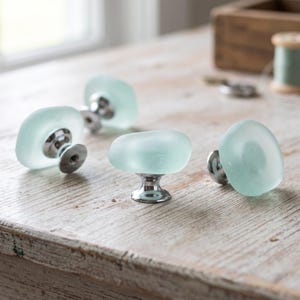May include: A set of four cabinet knobs featuring smooth, translucent, pale seafoam green glass tops with a rounded, pebble-like shape. Each knob is attached to a polished, reflective silver-toned metal base with a circular mounting plate.