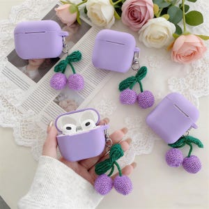 May include: Several lavender-colored AirPod cases with matching cherry charms. The cases are square and have a silver clasp. The charms are made of purple knitted cherries with green leaves. One case is open, revealing white earbuds.
