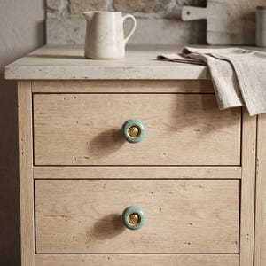 May include: A light-colored wooden cabinet featuring two drawers, each equipped with a round, speckled teal ceramic knob with a central brass-toned metal insert. The cabinet has a smooth, light-toned stone countertop surface.