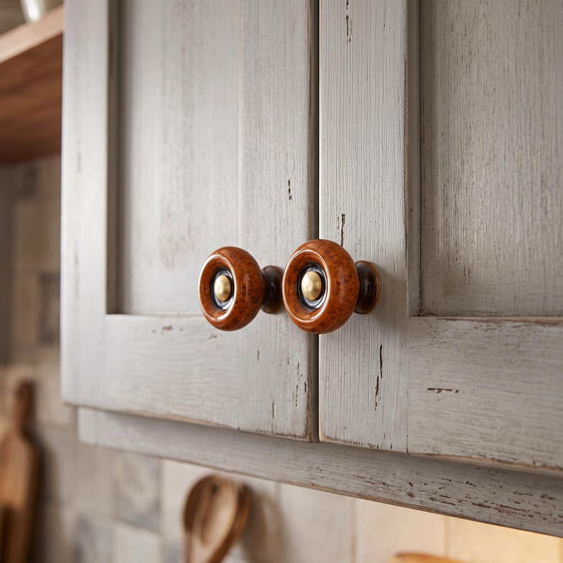 May include: Two round cabinet knobs featuring a glossy, mottled amber-brown ceramic outer ring, a dark brown inner ring, and a central brushed brass-toned metal button, mounted on light gray, distressed-finish wooden cabinet doors.