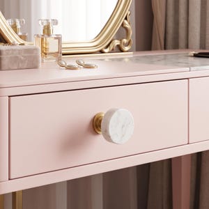 May include: A light pink vanity table featuring a single drawer with a round white marble knob and a gold-toned metal base. The tabletop surface is partially covered with a white marble inlay, and a gold-framed mirror sits on the back edge.
