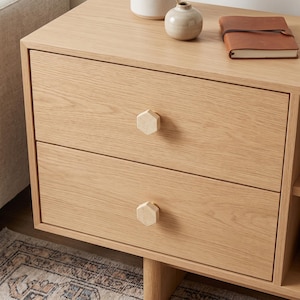 May include: A light oak wood bedside table featuring two drawers with hexagonal light-coloured knobs. On the top surface sits a small beige ceramic vase and a brown leather-bound notebook. The unit has a clean, natural wood grain finish throughout.