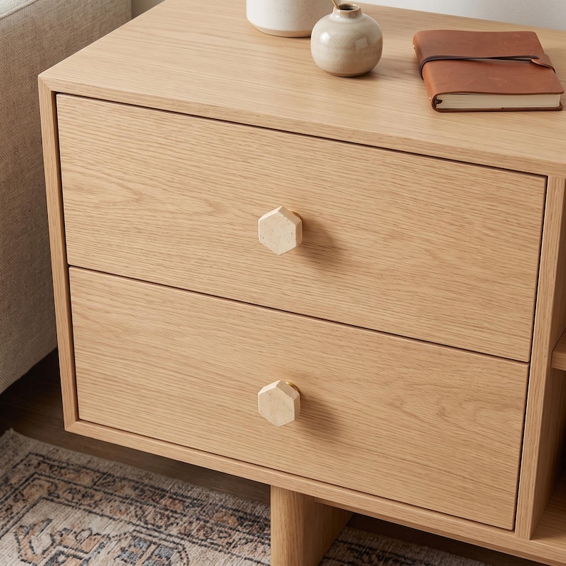 May include: A light oak wood bedside table featuring two drawers with hexagonal light-coloured knobs. On the top surface sits a small beige ceramic vase and a brown leather-bound notebook. The unit has a clean, natural wood grain finish throughout.