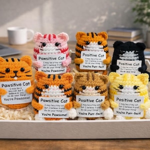 May include: A selection of handmade crochet cat figurines in various colours, including orange, pink, black, and yellow, each holding a sign with positive messages. The cats are displayed in a white tray.