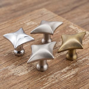 May include: Four metal cabinet knobs with a four-pointed star shape. The set includes one polished chrome knob, two brushed nickel knobs with a matte finish, and one antique brass knob. Each knob features a central stem and a rounded base.