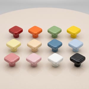 May include: A collection of twelve square cabinet knobs arranged in a grid, featuring a glossy finish in various solid colours including red, orange, green, yellow, pale yellow, cream, blue, light blue, pink, light pink, white, and black.