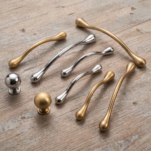 May include: A collection of cabinet hardware including two spherical knobs and six curved drawer pulls. The hardware features a mix of polished chrome and matte gold finishes, with each pull having rounded, teardrop-shaped ends.