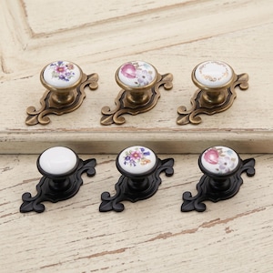 May include: A set of six drawer knobs featuring round white ceramic tops with floral patterns or gold filigree designs. Three knobs have antique brass-toned metal backplates, while the other three have matte black metal backplates.