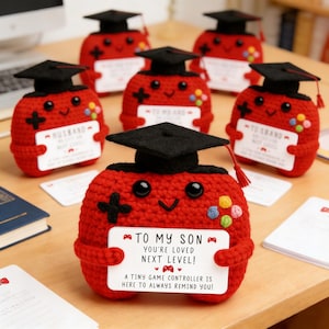 May include: Red crocheted game controller figurines wearing black graduation caps. Each controller holds a white sign with text like "TO MY SON" and "YOU'RE LOVED NEXT LEVEL!" Colorful buttons and a cross design are visible on the controllers.