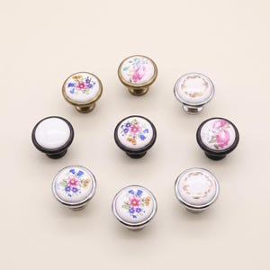 May include: A collection of eight round cabinet knobs featuring white ceramic centers. Some knobs display colorful floral patterns with pink, blue, and yellow flowers, while others feature gold-toned decorative accents and solid white surfaces.