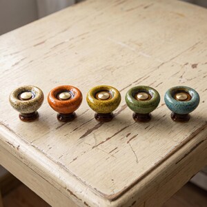 May include: Five round ceramic cabinet knobs with a glossy finish, each featuring a central metallic gold-toned disc. The knobs are colored in beige, orange, yellow, green, and blue, and each is mounted on a small, dark brown circular base.